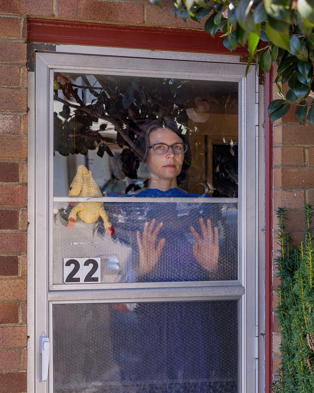 A woman in a blue dress looking through her screen door. There is a homemade, crocheted rubber chicken in the door as well.